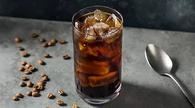 The image shows a glass of iced coffee, likely a cold brew, with ice cubes and coffee beans scattered on the surface next to it.