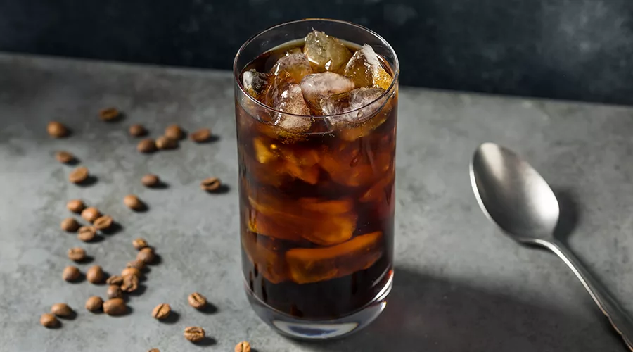 The image shows a glass of iced coffee, likely a cold brew, with ice cubes and coffee beans scattered on the surface next to it.
