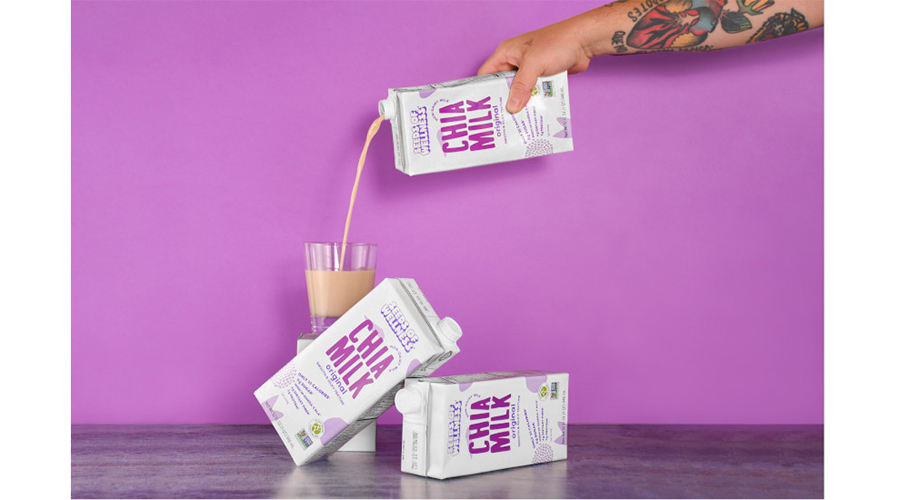 A tattooed arm pouring a carton of Benexia Seeds of Wellness Chia Milk in a glass, two cartons laying on their side, a purple wall in the background.
