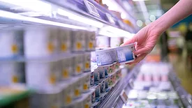 A woman's hand grabbing a yogurt pack from the shelf, the foreground and background are blurred.