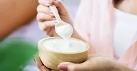A woman is holding a small wooden bowl of yogurt on one hand and a spoon with yogurt in the other. The background is blurred showing a cropped image of two people.