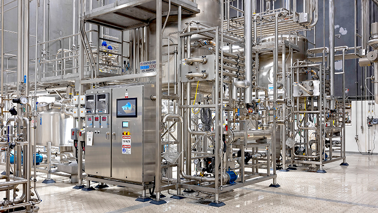Stainless steel industrial processing equipment with pipes, tanks, and control panels in a clean room.