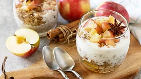 Dessert in a glass cup and two spoons on a wooden board, another glass and apples in the background.