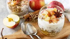 Dessert in a glass cup and two spoons on a wooden board, another glass and apples in the background.