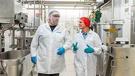 Two workers in a processing facility, in lab coats, hairnets, and gloves walking and talking to each other.