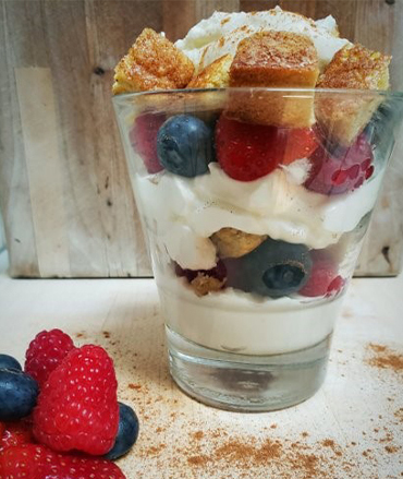 A yogurt parfait with French toast croutons and fresh fruit layers. More fruit and chocolate dusting are on the table.