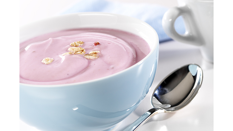 A bowl of yogurt on a table next to a spoon. A coffee cup and napkin in the background.