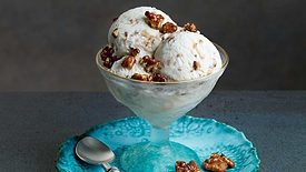 Scoops of buttercrunch ice cream with walnuts in an ice cream glass on a blue plate sitting on a dark gray table.  The plate contains two walnuts on one side and a spoon on the other.