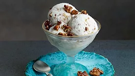 Scoops of buttercrunch ice cream with walnuts in an ice cream glass on a blue plate sitting on a dark gray table.  The plate contains two walnuts on one side and a spoon on the other.