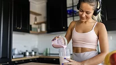 Woman in athleisure outfit pouring liquid yogurt into a container, while standing by the kitchen island.  The background is a blurred kitchen.