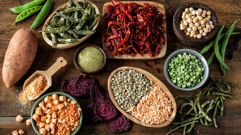 Overhead shot of bowls in various sizes and shapes containing dried peppers, herbs and spices, dried and fresh potatoes and green beans on the wooden table.