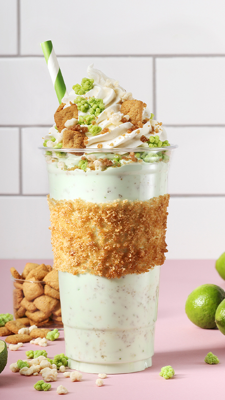 A keylime milkshake with inclusions on a pink counter with limes, cookies and crunchy white and green bits on a pink counter with white subway tiles in the background.