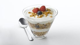 A yogurt parfait with layers of granola, blueberries and raspberries, with a spoon on the table on the left. The background is white.
