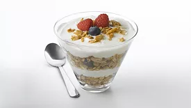 A yogurt parfait with layers of granola, blueberries and raspberries, with a spoon on the table on the left. The background is white.