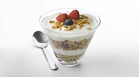 A yogurt parfait with layers of granola, blueberries and raspberries, with a spoon on the table on the left. The background is white.