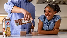 A dad (head is cropped out) is pouring chocolate milk into glasses in the kitchen, his daughter is beside him smiling.