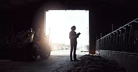A young woman standing inside a barn silhouetted by the light of the open barn door. She's holding a clipboard.