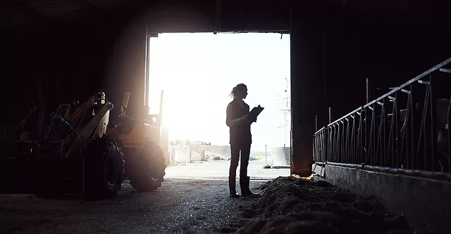 A young woman standing inside a barn silhouetted by the light of the open barn door. She's holding a clipboard.