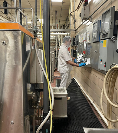 A worker in protective gear operates machinery and control panels in a busy industrial food processing plant.