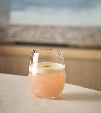 A short glass with a pink beverage, white foam, and an orange garnish on a light tablecloth.