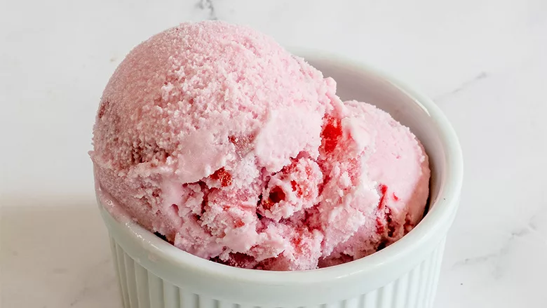 Two scoops of pink strawberry ice cream in a white ceramic ramekin on a white marble surface.
