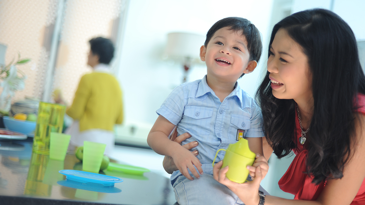 A young Asian mother holding her son and a sippy cup, an older woman is in the blurred background.