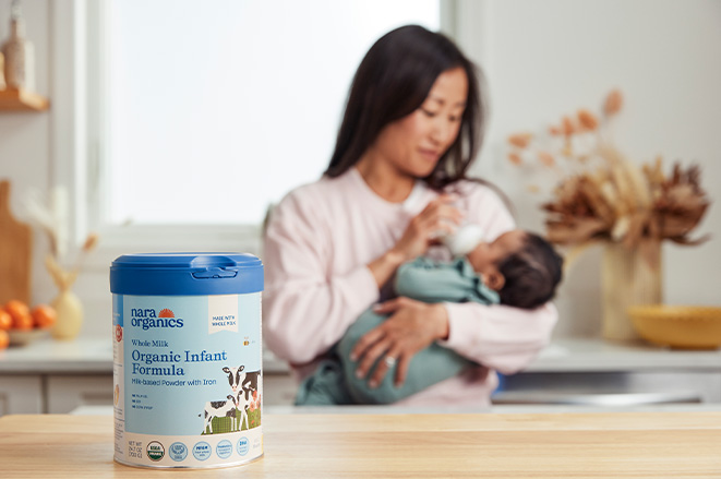 A tub of Nara Organics Whole Milk Infant Formula on the counter, a young Asian mother holding and feeding her infant in the blurred background.