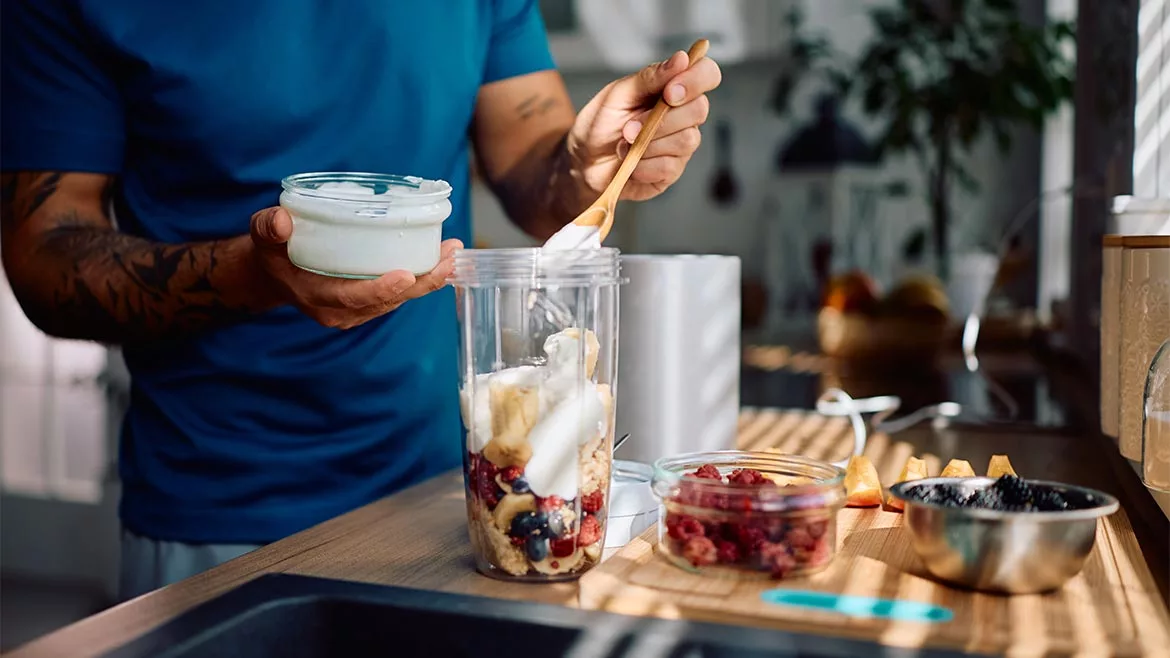 Close up of man adding Greek yogurt while preparing healthy smoothie in the kitchen.