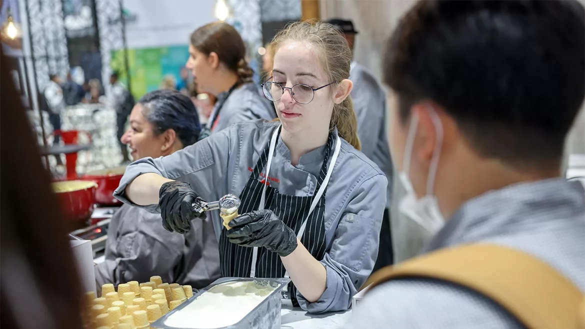 Young woman serving ice cream on a cone surrounded by other workers.