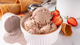 A bowl if ice cream with strawberry slices, ice cream cones in the background and a spilled over container of protein powder.