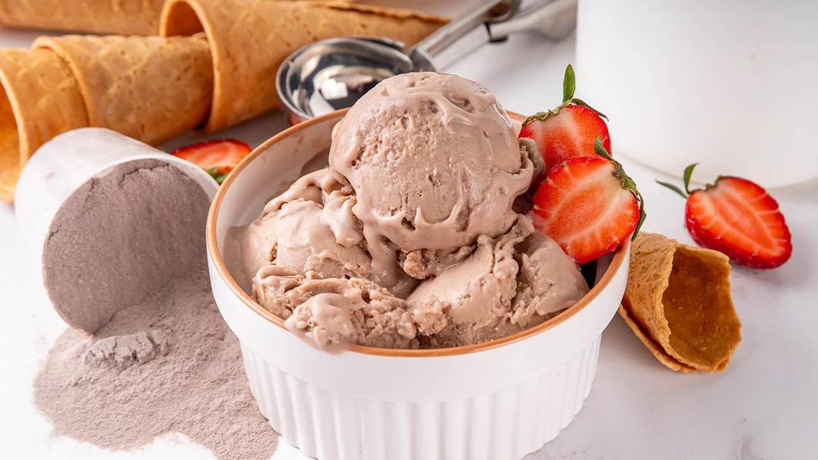 A bowl if ice cream with strawberry slices, ice cream cones in the background and a spilled over container of protein powder.