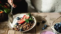  Female hand using a spoon to eat vanilla yogurt with chocolate granola and berry fruits in plate over wooden table.