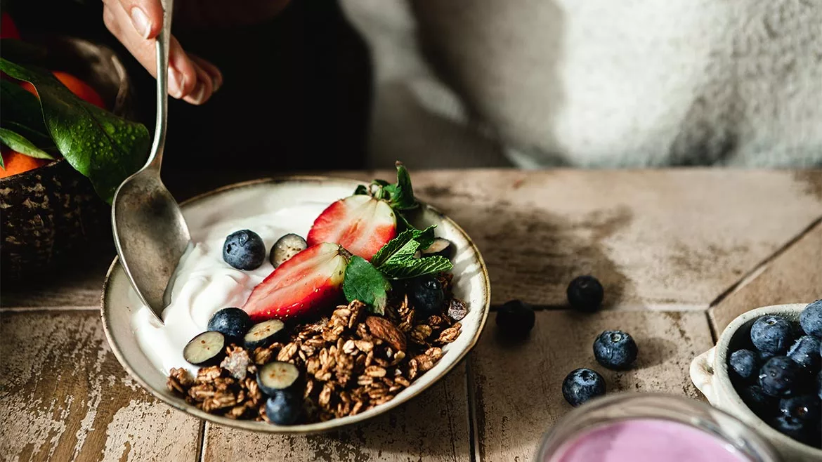  Female hand using a spoon to eat vanilla yogurt with chocolate granola and berry fruits in plate over wooden table.