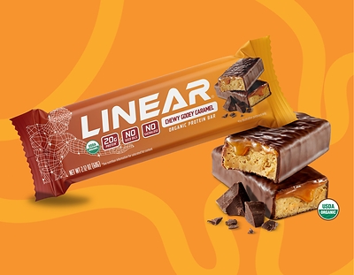 A Linear Chewy Gooey Caramel bar, one side laying on an opened bar broken in half, a yellow and orange patterned background.
