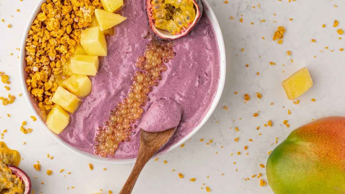Smoothie bowl with sprinkles and fruit Close-up of a vibrant purple smoothie bowl topped with pineapple chunks, granola, passion fruit, and fruit pearls, with a wooden spoon resting in the bowl and a fresh mango on a light marble surface.