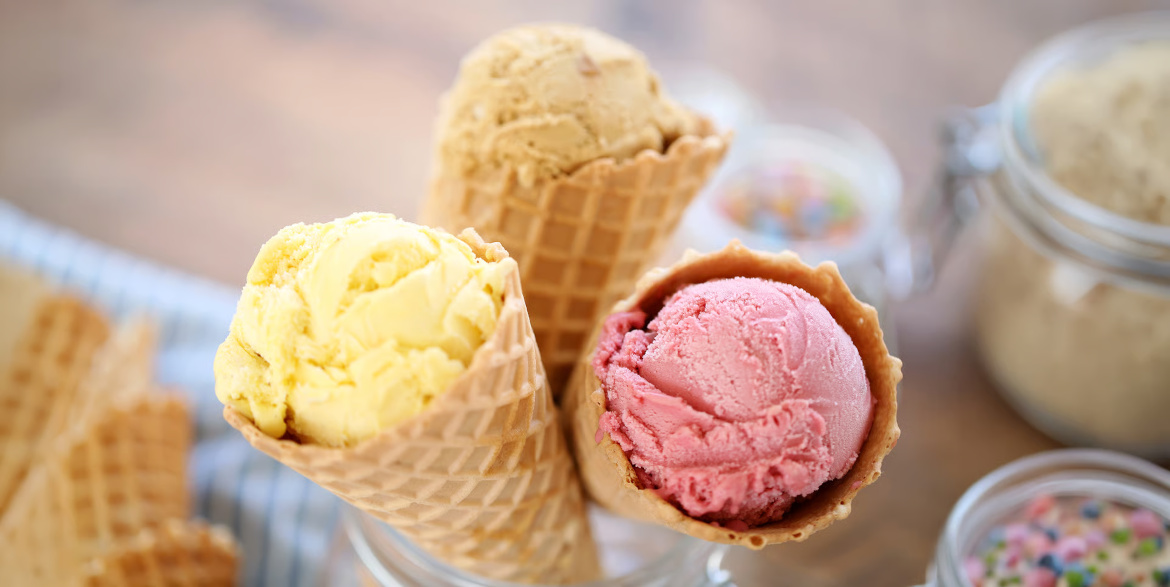 Three different flavored ice cream scoops in cones with a blurred background or extra cones and jars of sprinkles.