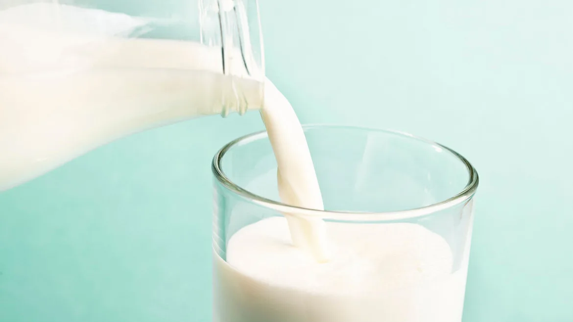 Milk being poured from a glass container into a glass, the background is a teal color.
