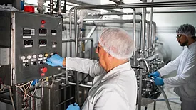 Two workers in protective clothing monitor and operate machinery in a modern dairy processing plant, showcasing hygiene and production processes.