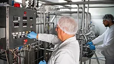 Two workers in protective clothing monitor and operate machinery in a modern dairy processing plant, showcasing hygiene and production processes.